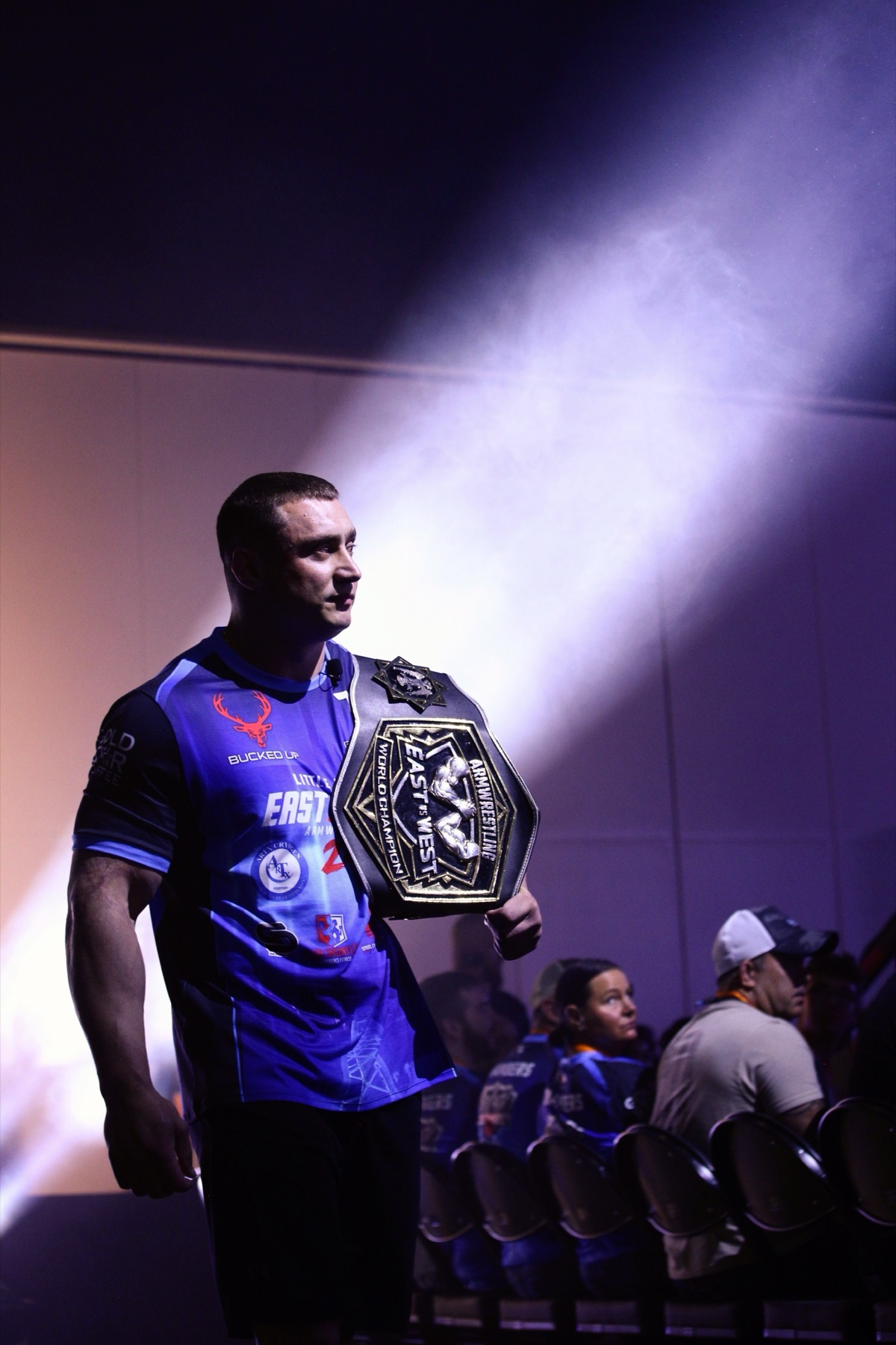 East vs West 24: an armwrestling world champion walks out through the haze holding the title belt